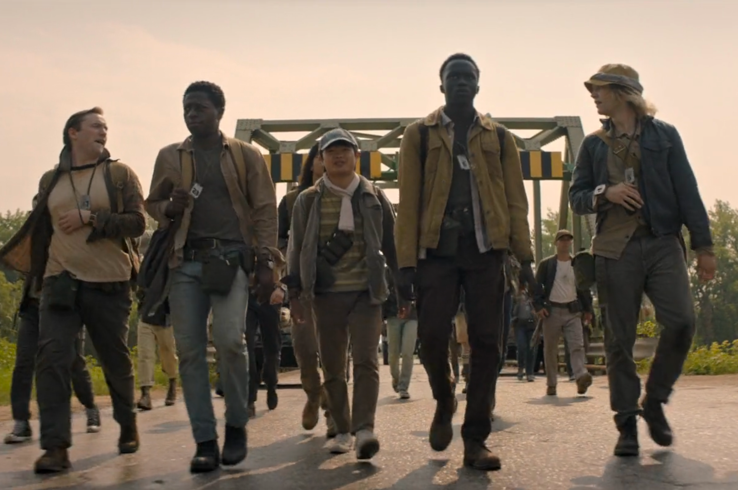 Group of teenage boys walking down a desolate road under a gray sky, soldiers in the distance—scene from The Long Walk (2025) film adaptation.